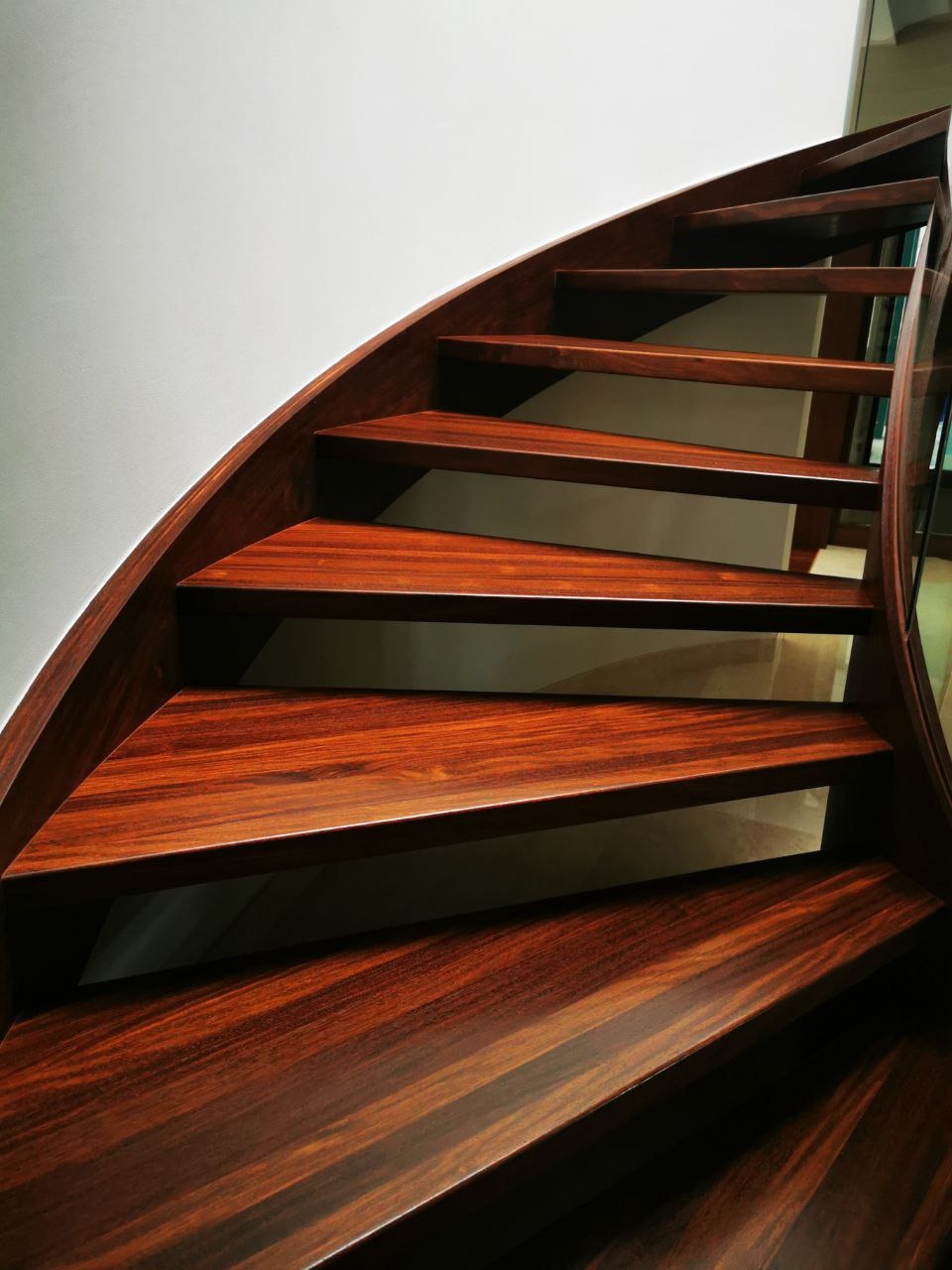Polerowane, mahoniowe schody kręcone z drewnianą poręczą i szklaną balustradą, widok od dołu.