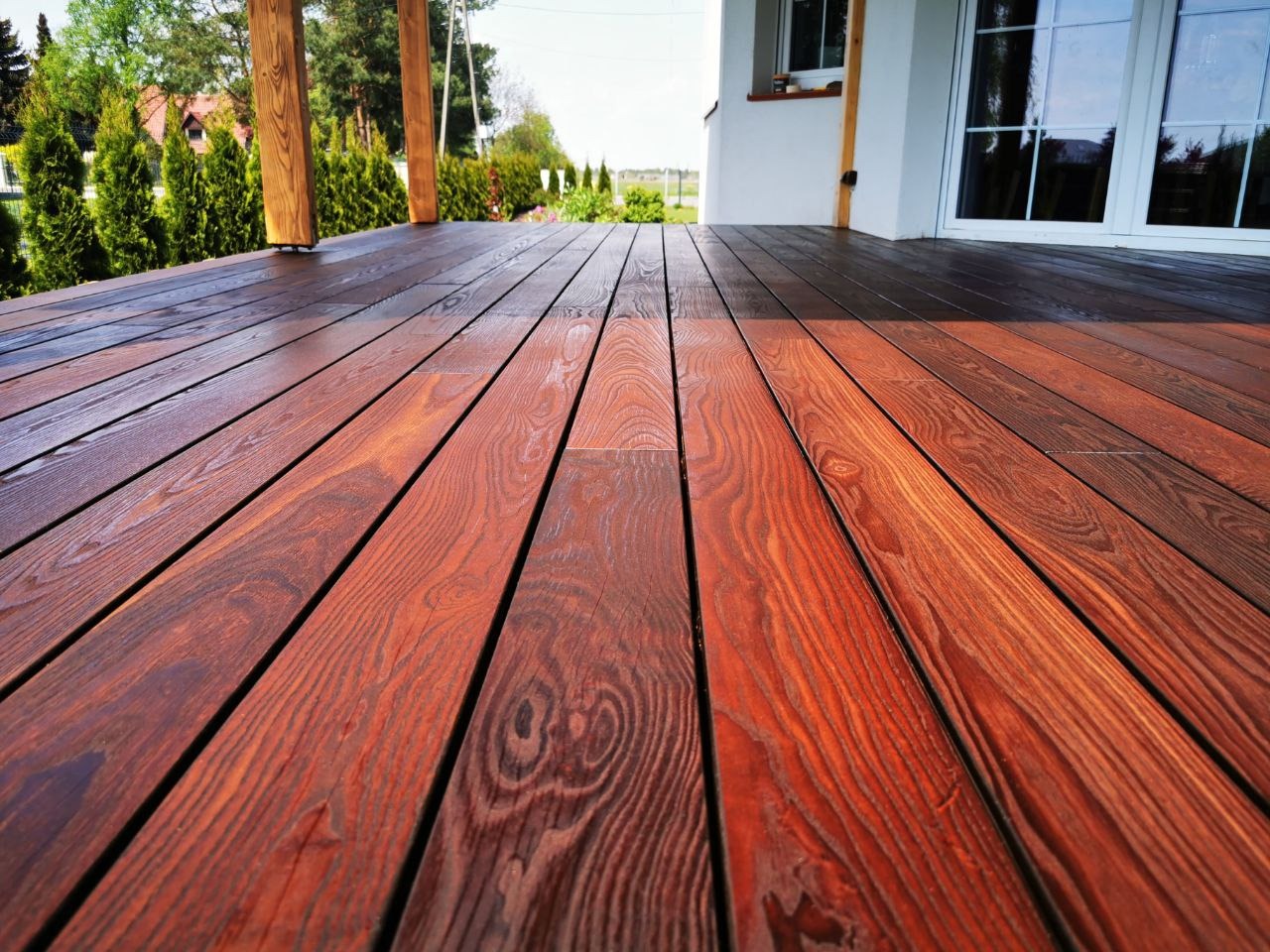 Świeżo polakierowany taras z desek o wyraźnym usłojeniu w odcieniach brązu i mahoniu, z widoczną fakturą drewna, prowadzący do białego domu z dużymi oknami.