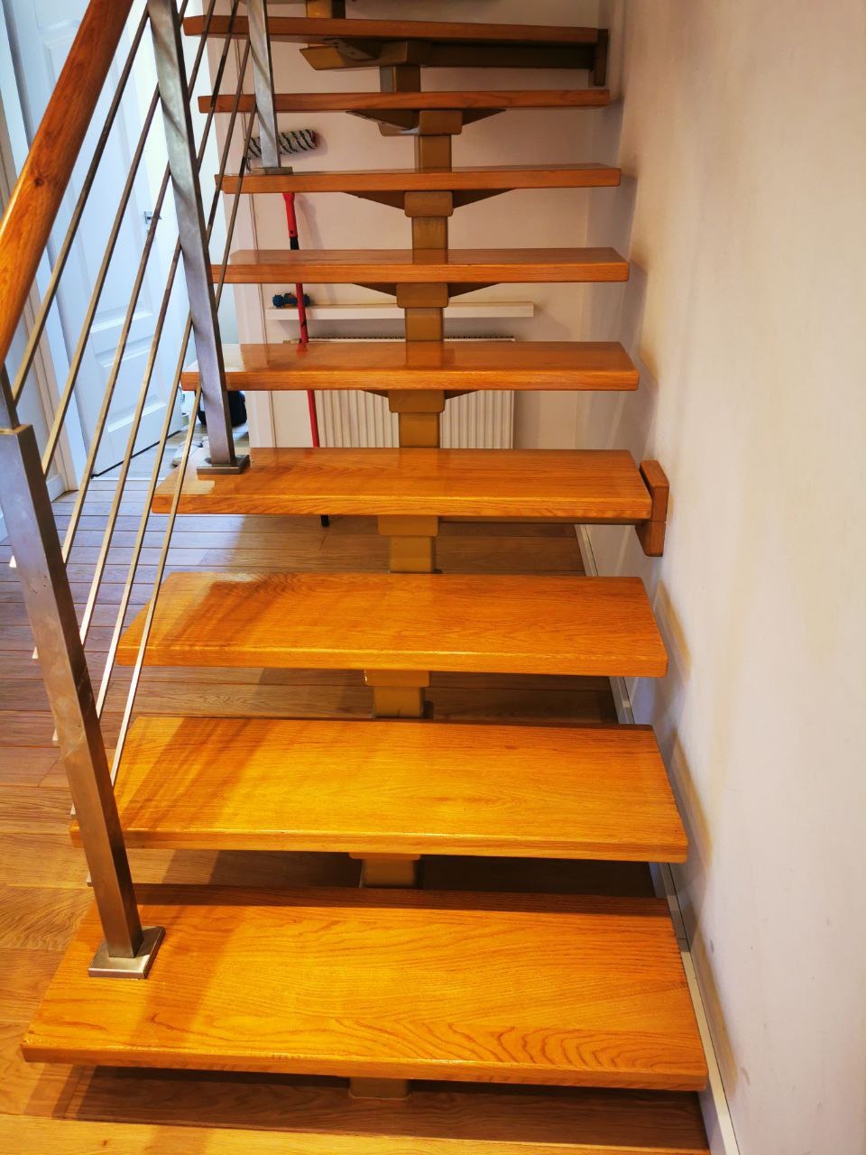 Drewniane schody z metalową balustradą, widok z dołu, naturalne oświetlenie podkreśla usłojenie drewna, w tle biała ściana i fragment podłogi z paneli.