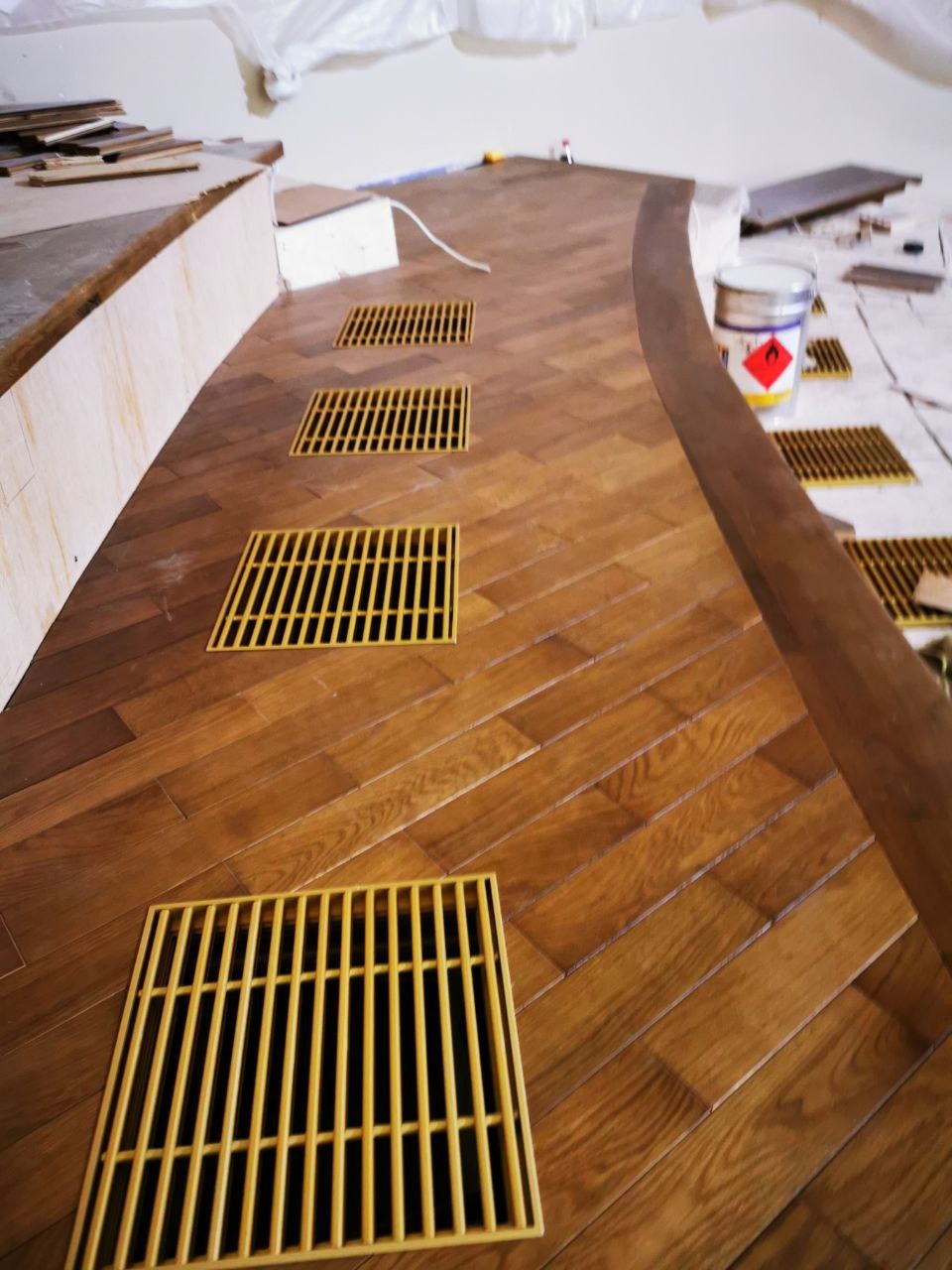 Ułożony parkiet w jodełkę z wmontowanymi kratkami wentylacyjnymi, widoczne wiadro z lakierem i przygotowane deski podłogowe.