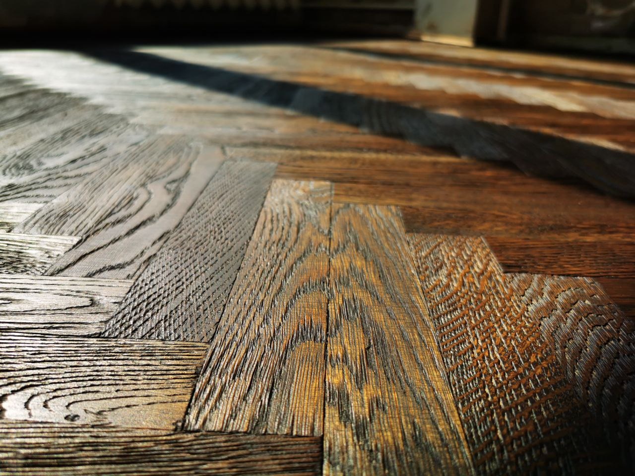 Drewniana podłoga ułożona w jodełkę, z widocznym usłojeniem i refleksami światła, ukazująca detale wykończenia.