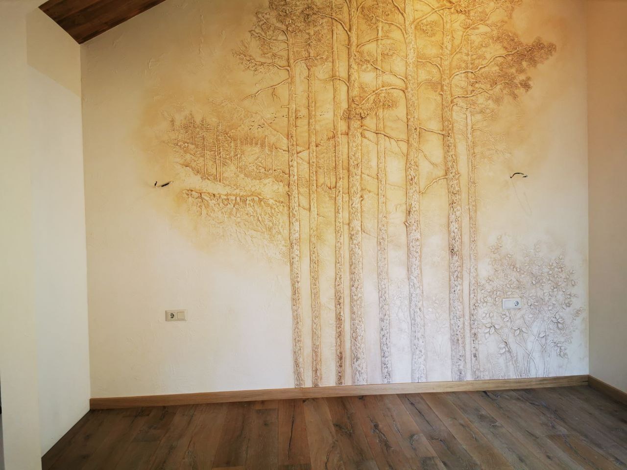 Ściana w pokoju z dekoracyjnym szpachlowaniem przedstawiającym motyw lasu: wysokie drzewa i krzewy, brązowe panele podłogowe, widoczne gniazdka elektryczne.