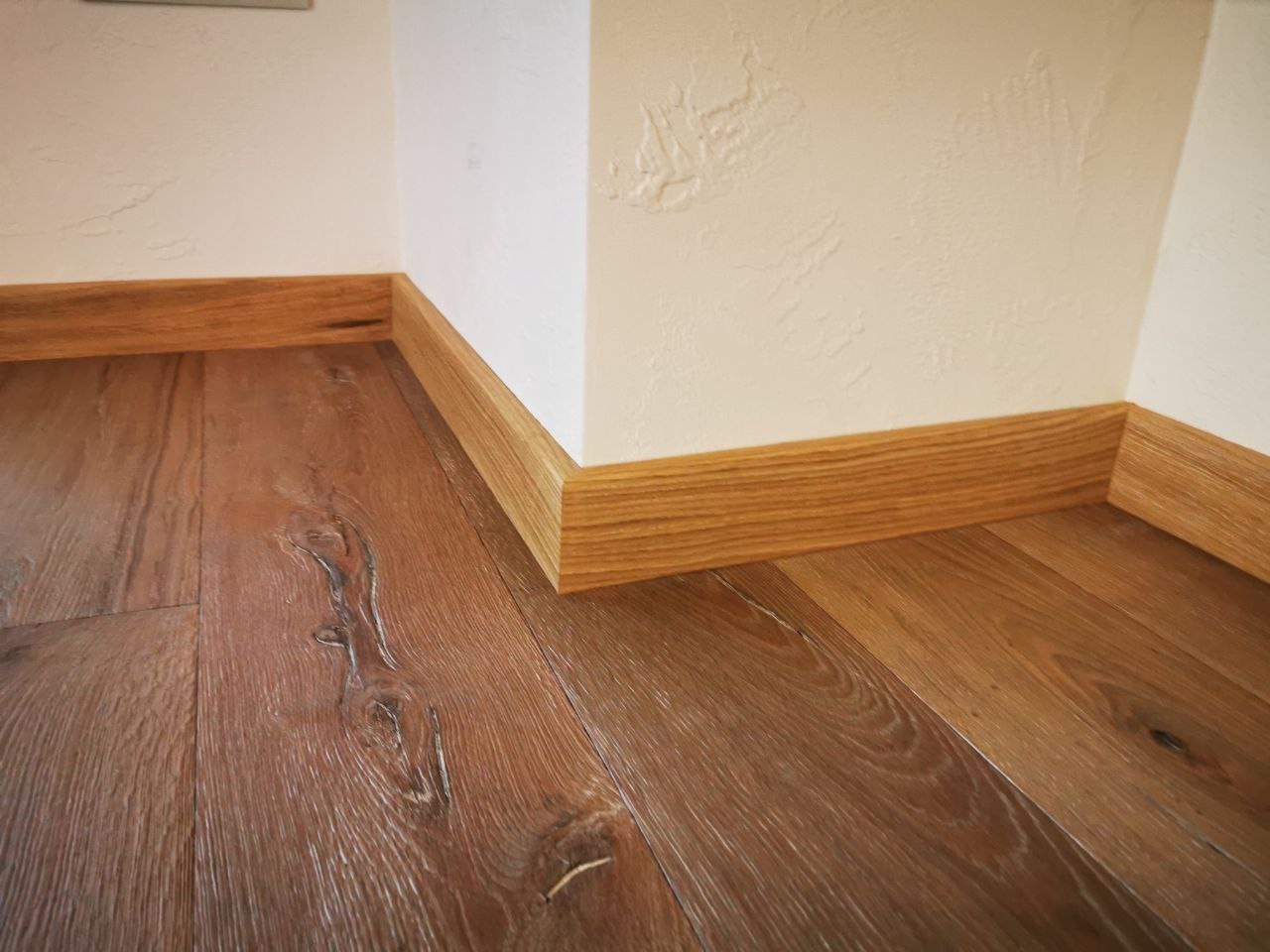 Fragment podłogi z ciemnego drewna z widocznymi słojami, wykończonej jasnymi listwami przypodłogowymi przy białej ścianie z fakturą.