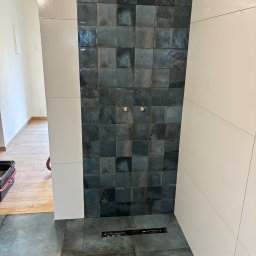 MWB USŁUGI OGÓLNOBUDOWLANE Mateusz Woś - Ściana prysznicowa wyłożona ciemnymi, kwadratowymi płytkami z efektem postarzenia, sąsiadująca z białą ścianą wyłożoną dużymi, prostokątnymi płytami; widoczny odpływ liniowy w podłodze.