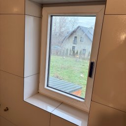 MWB USŁUGI OGÓLNOBUDOWLANE Mateusz Woś - Jasne, błyszczące płytki ceramiczne ułożone wokół okna w łazience, widok na zewnątrz na dom i ogród.