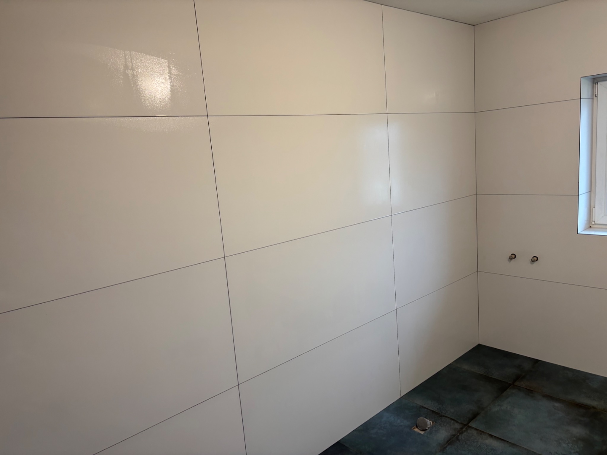 Wnętrze łazienki po remoncie, ściany wyłożone jasnymi, błyszczącymi płytami wielkoformatowymi z cienkimi fugami, ciemna podłoga z płytek imitujących kamień, widoczne przyłącza wodne i fragment okna.