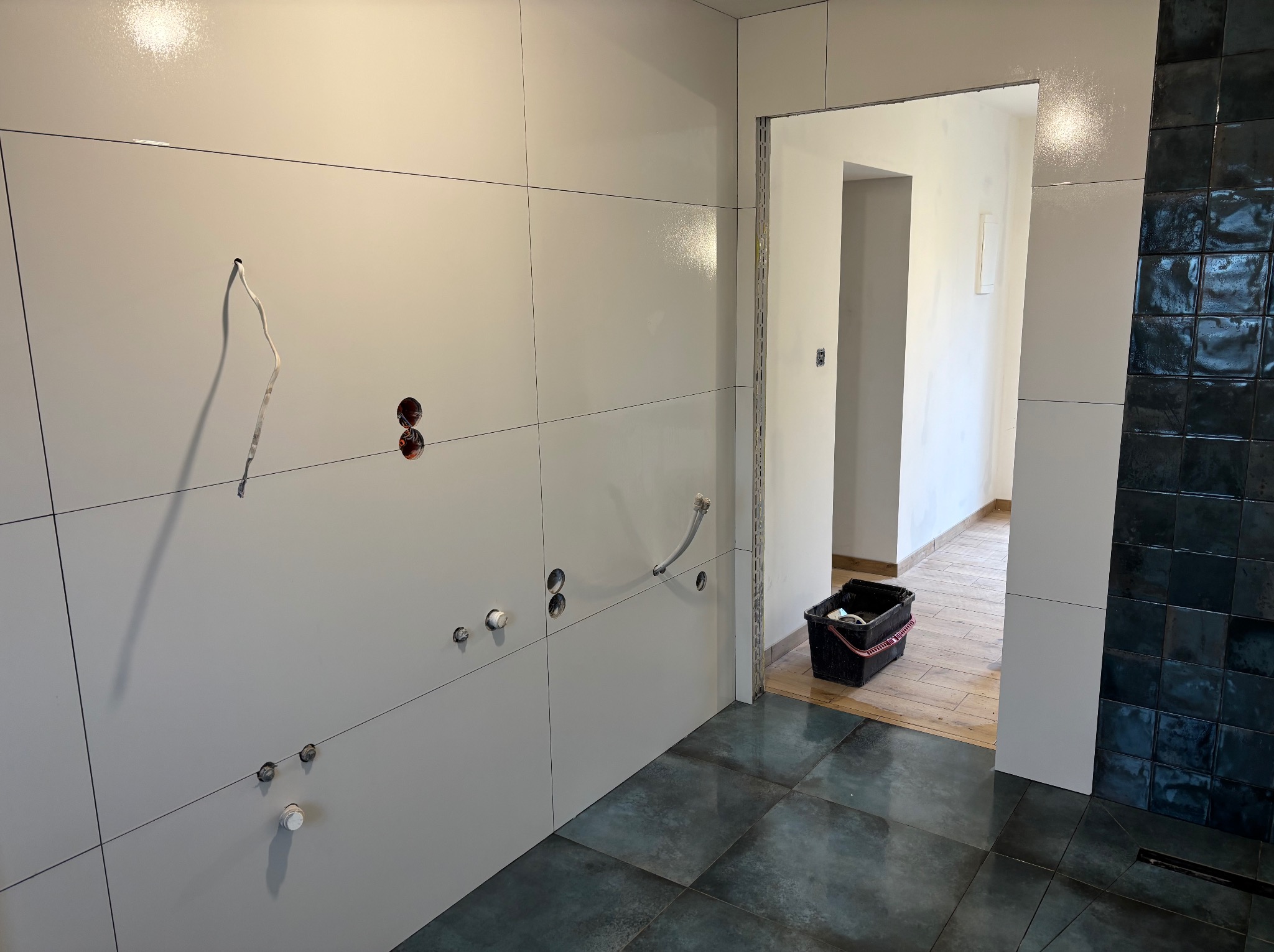 Łazienka w trakcie prac wykończeniowych: białe, wielkoformatowe płytki ścienne z widocznymi instalacjami, ciemna, kwadratowa mozaika na ścianie oraz ciemne płytki podłogowe, wiadro z narzędziami...