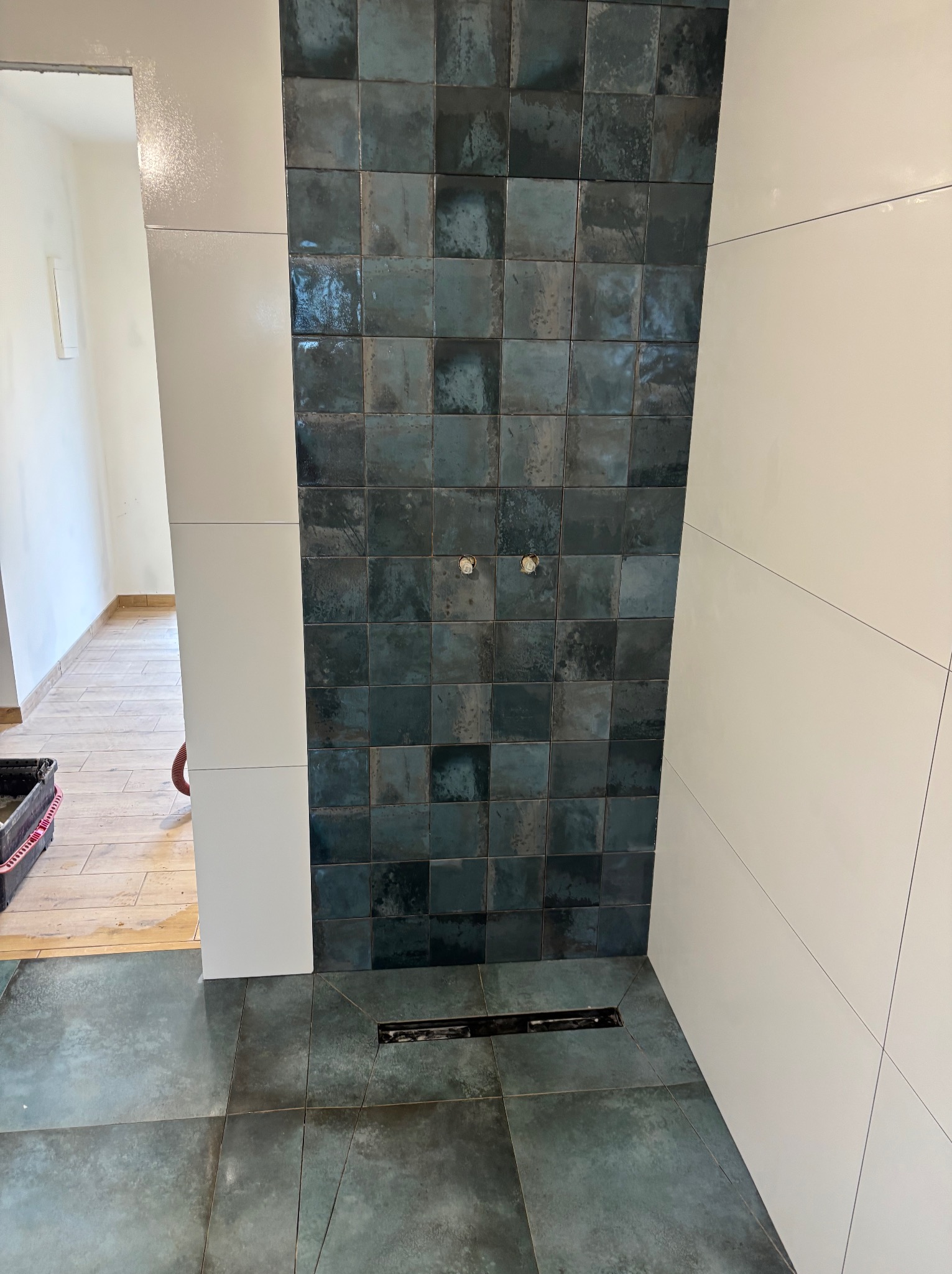 Ściana prysznicowa wyłożona ciemnymi, kwadratowymi płytkami z efektem postarzenia, sąsiadująca z białą ścianą wyłożoną dużymi, prostokątnymi płytami; widoczny odpływ liniowy w podłodze.