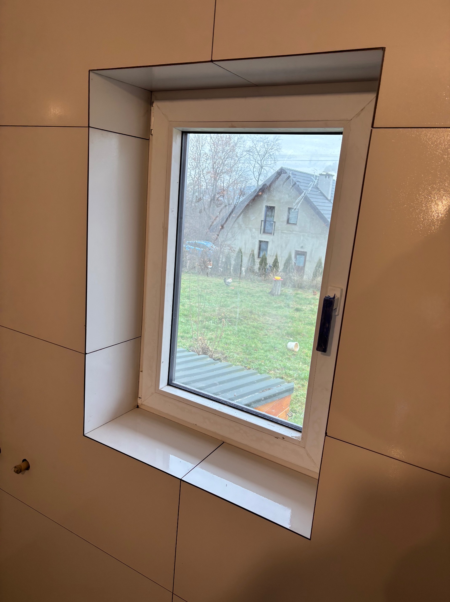 Jasne, błyszczące płytki ceramiczne ułożone wokół okna w łazience, widok na zewnątrz na dom i ogród.