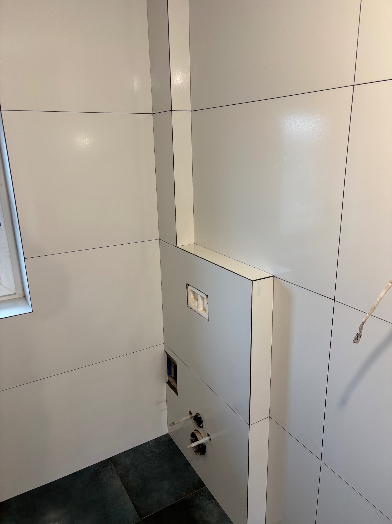 Świeżo ułożone, błyszczące, białe płytki ścienne z czarnymi fugami w łazience, widoczna zabudowa stelaża podtynkowego WC z przyłączami hydraulicznymi, czarne płytki podłogowe.