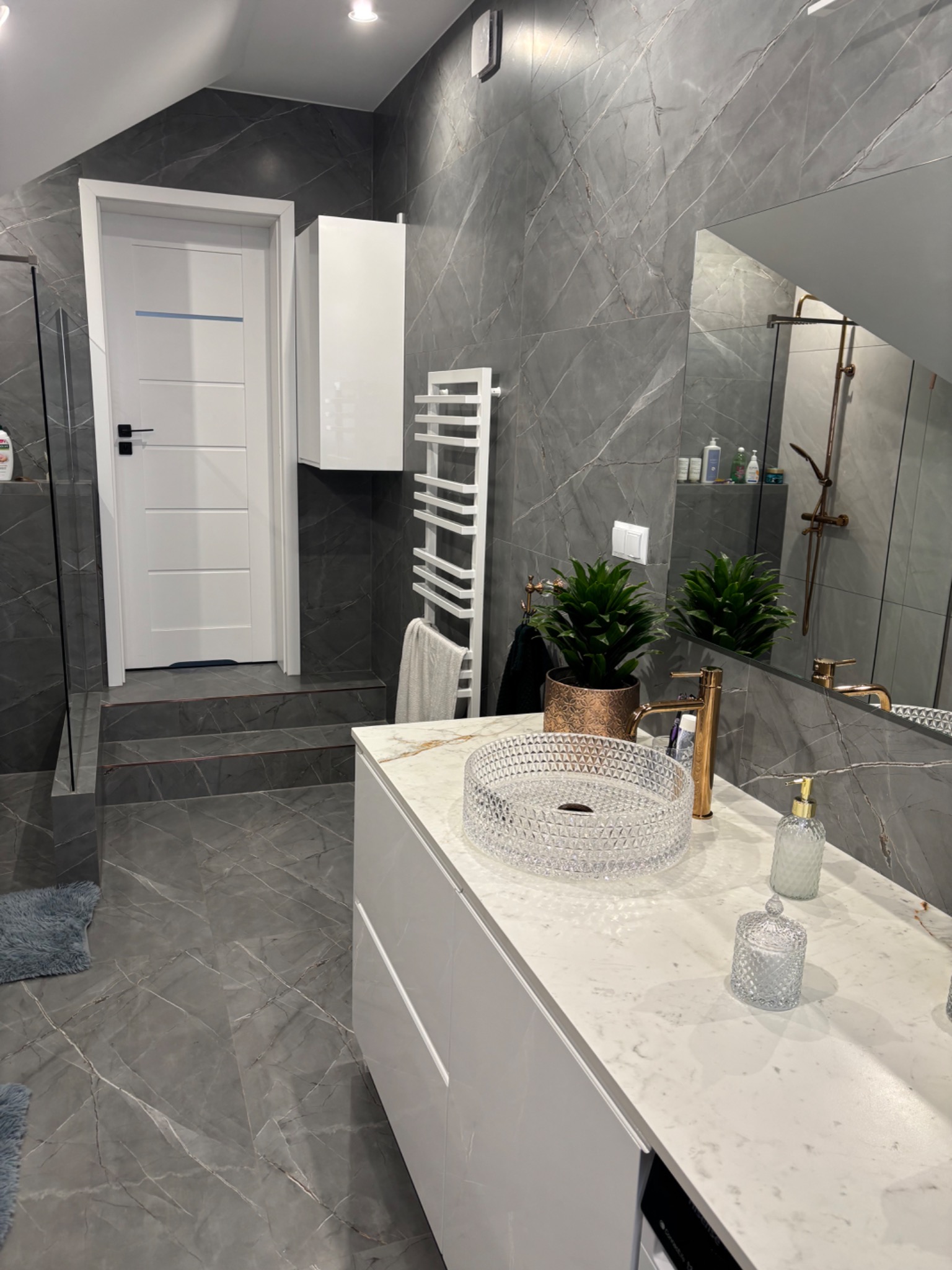 Nowoczesna łazienka z szarymi płytkami imitującymi marmur, białą szafką z umywalką z kryształowego szkła, złotą baterią i dekoracyjnymi roślinami w doniczkach.