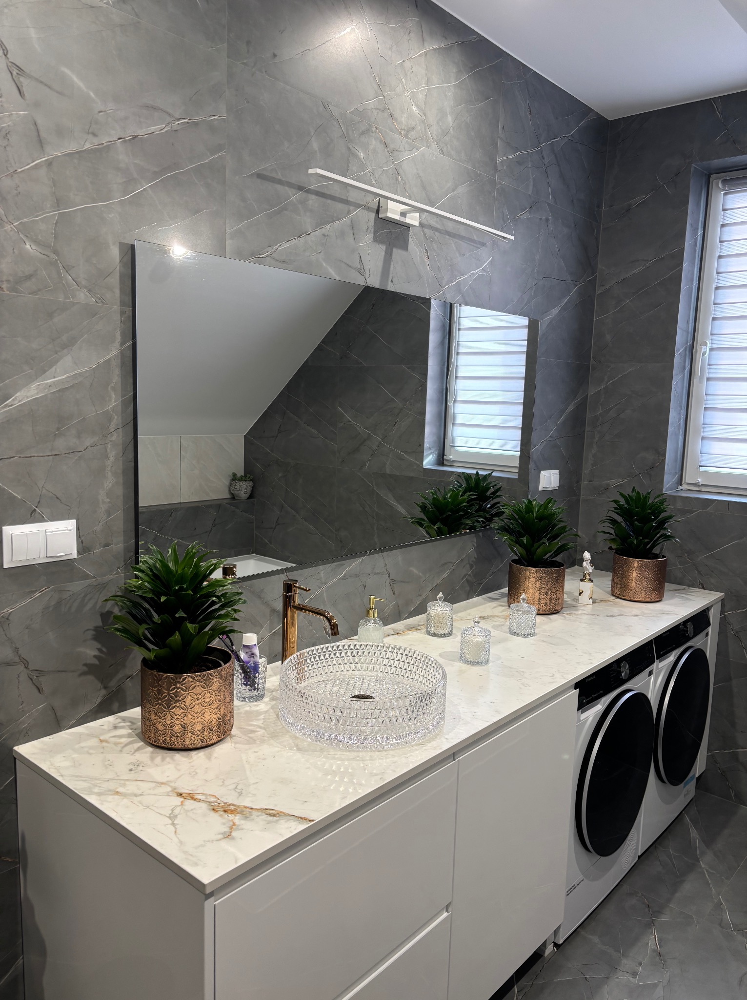 Nowoczesna łazienka z szarymi marmurowymi płytkami, umywalką kryształową na blacie z białego marmuru, złotą armaturą i dwoma pralkami z czarnymi okrągłymi drzwiami w zabudowie.