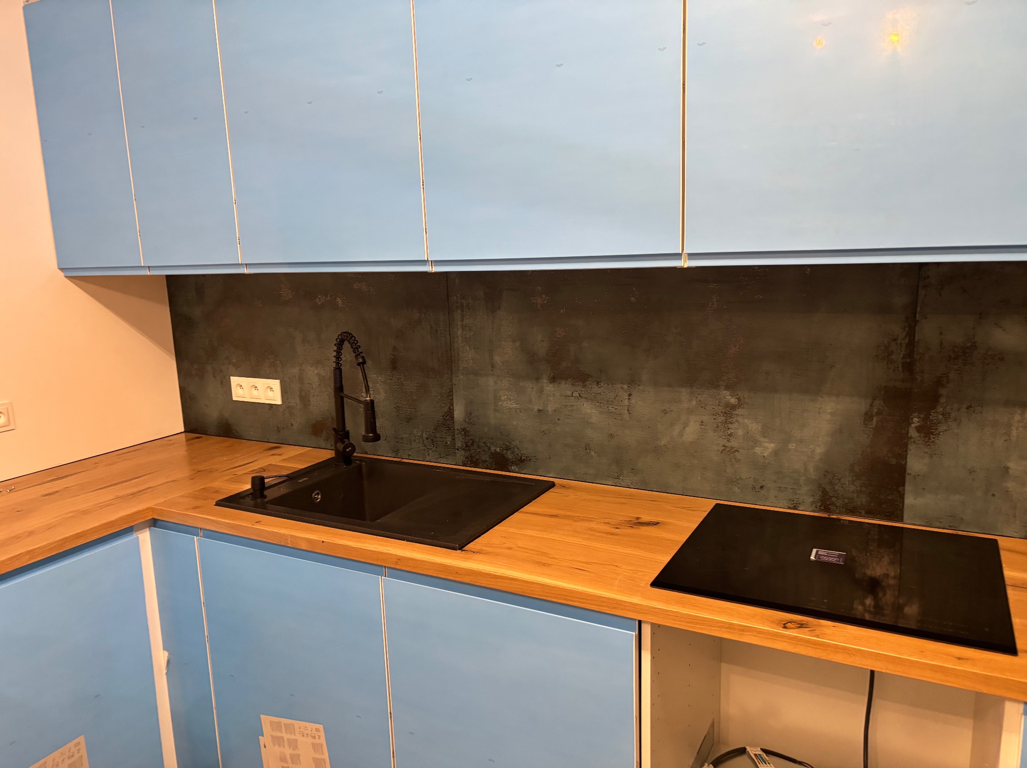 Nowoczesna kuchnia z błękitnymi szafkami, drewnianym blatem i czarnym zlewem z baterią, uzupełniona płytą indukcyjną i ciemną ścianą nad blatem.