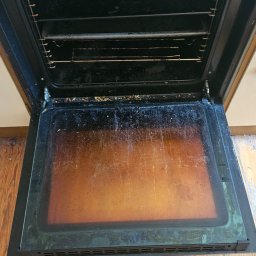 SPRZĄTANIE NIERUCHOMOŚCI MAŁGORZATA KUFERA - Wnętrze bardzo zabrudzonego piekarnika z otwartymi drzwiczkami, widoczne przypalone resztki jedzenia na dnie i ściankach, ruszt brudny, na podłodze drewniana podłoga z widocznymi śladami użytkowania.