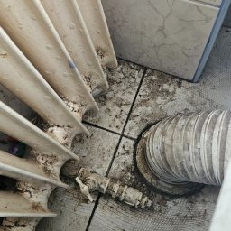 SPRZĄTANIE NIERUCHOMOŚCI MAŁGORZATA KUFERA - Zanieczyszczony grzejnik żeberkowy z brudnymi rurami i zaworem termostatycznym na brudnej podłodze wyłożonej płytkami ceramicznymi.