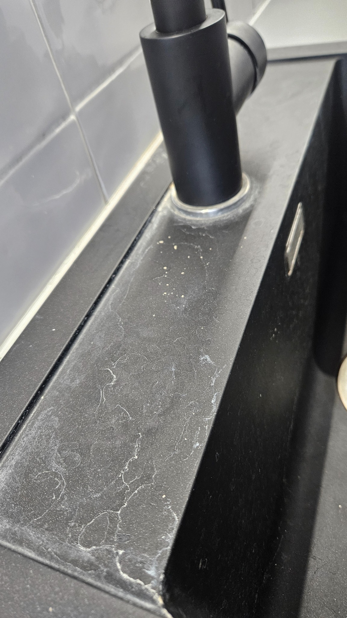 Czarny zlew kuchenny z widocznymi śladami kamienia i zaciekami wodnymi na powierzchni, czarna bateria. Widoczne szare płytki na ścianie.