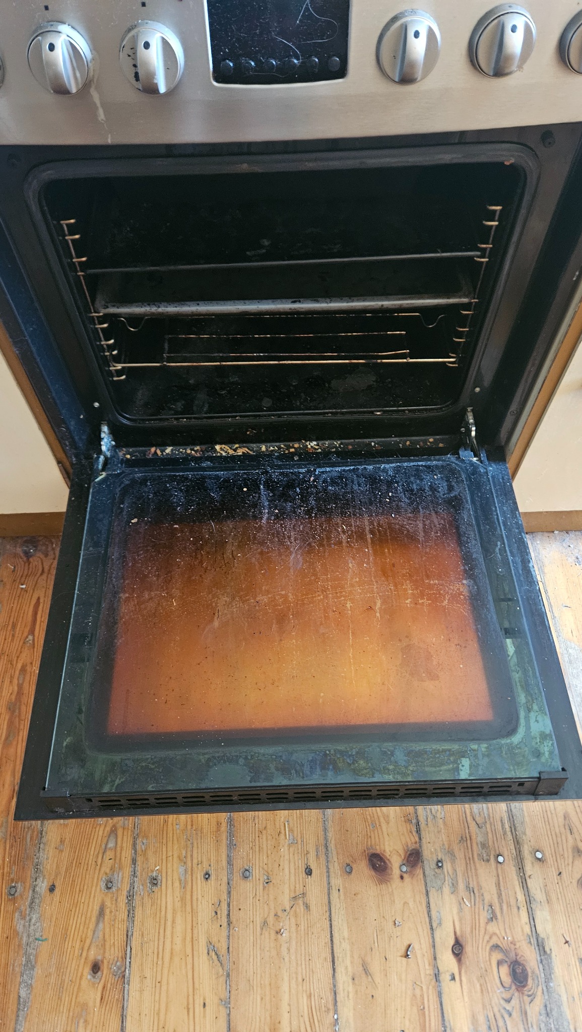 Wnętrze bardzo zabrudzonego piekarnika z otwartymi drzwiczkami, widoczne przypalone resztki jedzenia na dnie i ściankach, ruszt brudny, na podłodze drewniana podłoga z widocznymi śladami użytkowania.