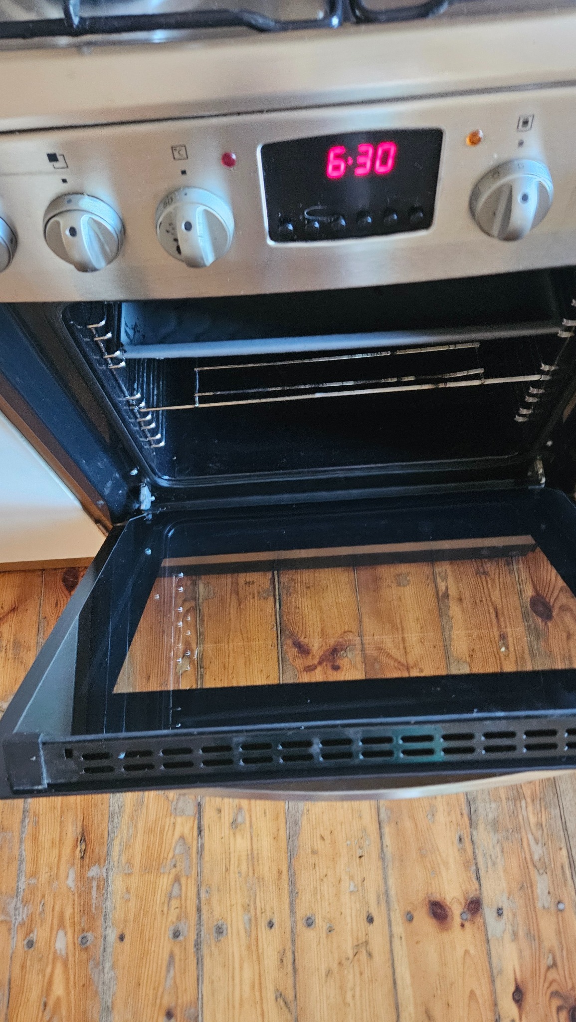 Wnętrze otwartego piekarnika ze srebrną obudową, widoczny zegar cyfrowy wskazujący godzinę 6:30, ruszt i drewniana podłoga z widocznymi śladami użytkowania.