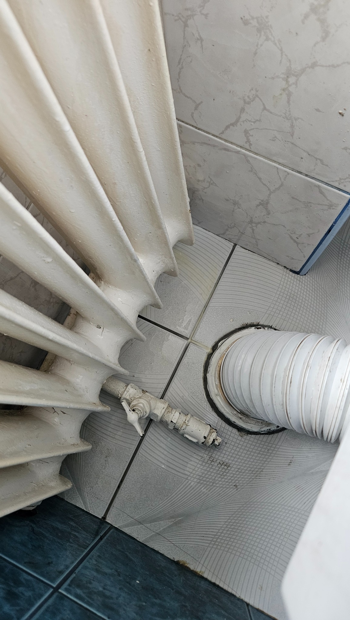 Widok z góry na brudny, biały grzejnik żeliwny z zaworem, białą rurę odpływową oraz płytki podłogowe z widocznymi zabrudzeniami i plamami, wymagające czyszczenia.