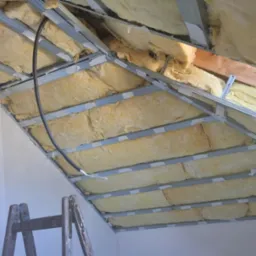 Skośny sufit poddasza w trakcie izolacji wełną mineralną, widoczne metalowe profile konstrukcyjne i częściowo otynkowana ściana oraz fragment drewnianej drabiny.