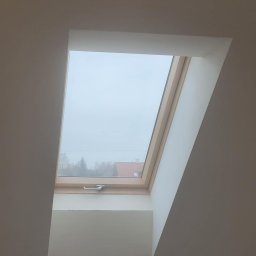 MARBUD - Zamontowane okno dachowe z drewnianą ramą, widok na zewnątrz w pochmurny dzień, widoczne drzewa i dach domu w oddali, gładkie białe wykończenie wnęki okiennej.