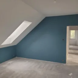 Pokój na poddaszu po szpachlowaniu, z pomalowaną na niebiesko ścianą do połowy wysokości, surową betonową podłogą i widocznym oknem dachowym oraz przejściem do kolejnego pomieszczenia.