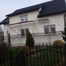 MARBUD - Dom jednorodzinny w trakcie prac elewacyjnych, widoczne rusztowania, okna zabezpieczone folią, świeżo nałożony tynk w kolorze złamanej bieli, czarny dach z dachówki ceramicznej, metalowe ogrodzenie...