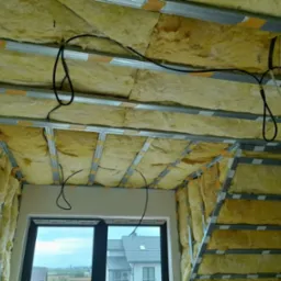 Skośny sufit poddasza w trakcie izolacji wełną mineralną, widoczne metalowe profile i przewody elektryczne, okno dachowe z widokiem na zabudowę jednorodzinną.