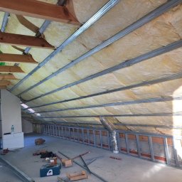 MARBUD - Poddasze w trakcie izolacji: widoczne metalowe profile konstrukcyjne, wełna mineralna między nimi, fragment okna z sylwetką osoby, narzędzia i materiały budowlane na podłodze.