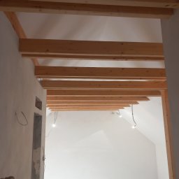 MARBUD - Wnętrze w trakcie prac wykończeniowych: drewniane belki stropowe, surowe ściany, widoczne przewody elektryczne. Ujęcie z dołu, perspektywa podkreśla wysokość pomieszczenia.