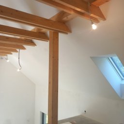 MARBUD - Poddasze z drewnianą konstrukcją stropu i oknem dachowym. Widoczne przygotowania do prac wykończeniowych, surowy beton na podłodze, białe ściany i elementy instalacji.