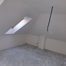 MARBUD - Poddasze po szpachlowaniu, przygotowane do dalszych prac wykończeniowych. Widoczne okno dachowe i wystające przewody elektryczne. Szare podłoże.