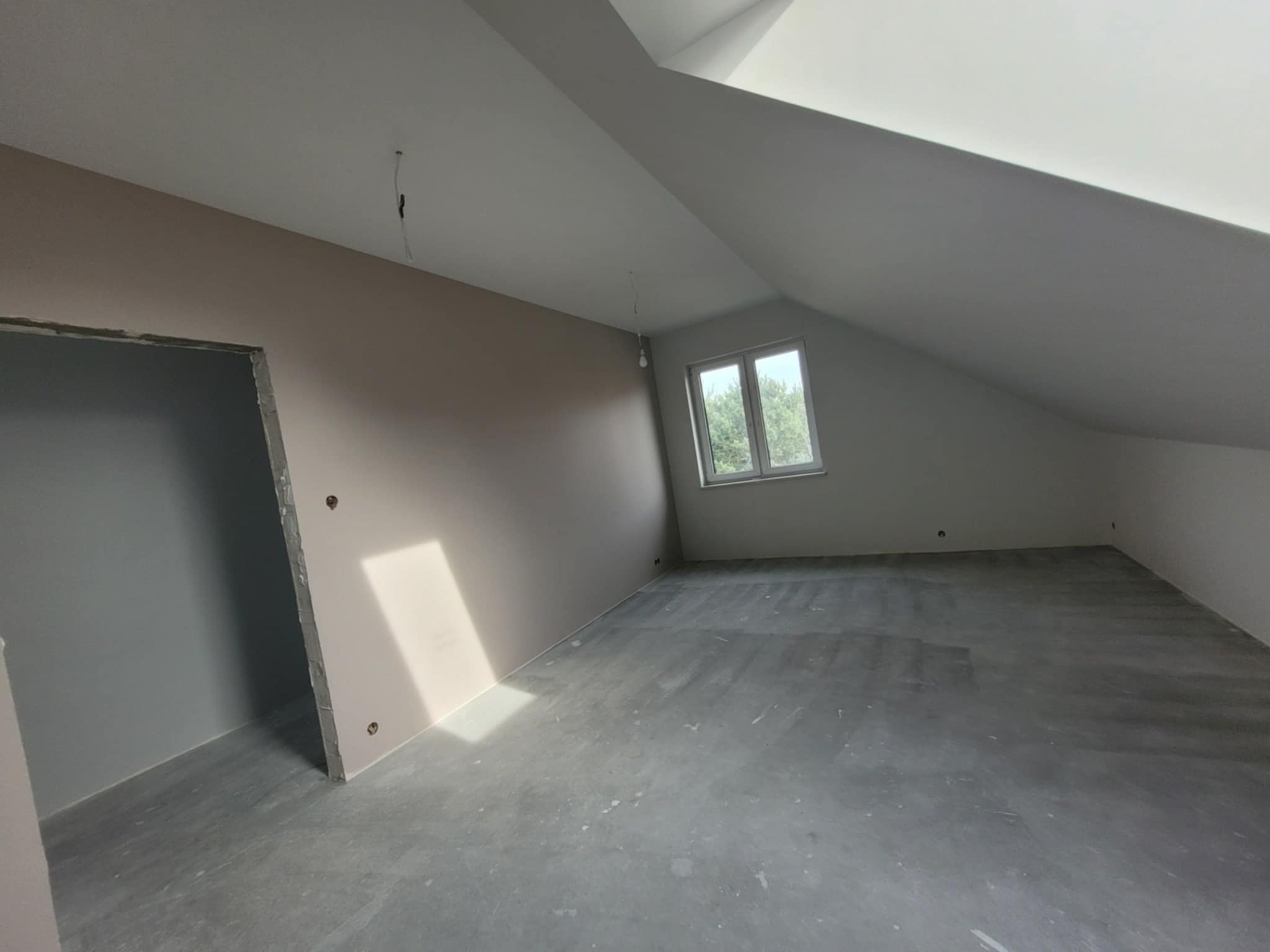 Pokój na poddaszu w trakcie prac wykończeniowych, widoczne ściany pomalowane na beżowo, surowa podłoga, okno z widokiem na drzewa i wystający kabel z sufitu.