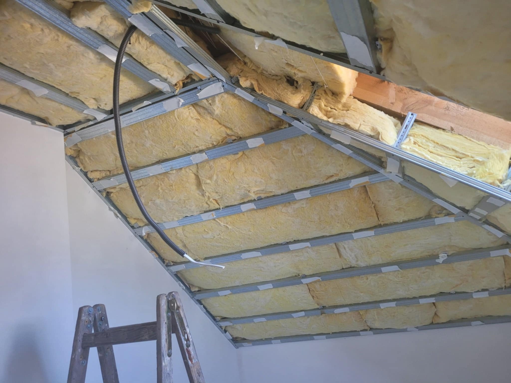 Skośny sufit poddasza w trakcie izolacji wełną mineralną, widoczne metalowe profile konstrukcyjne i częściowo otynkowana ściana oraz fragment drewnianej drabiny.