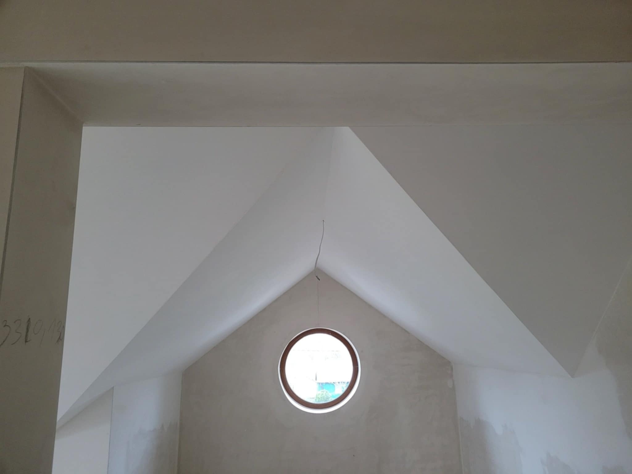 Wnętrze poddasza ze skośnym sufitem i okrągłym oknem, ściany w trakcie wykańczania, widoczne ślady po szpachlowaniu, sufit pomalowany na biało.