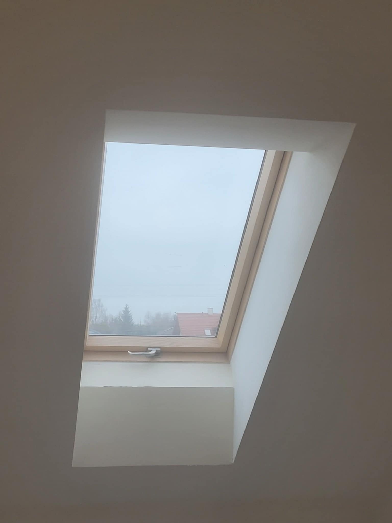 Zamontowane okno dachowe z drewnianą ramą, widok na zewnątrz w pochmurny dzień, widoczne drzewa i dach domu w oddali, gładkie białe wykończenie wnęki okiennej.