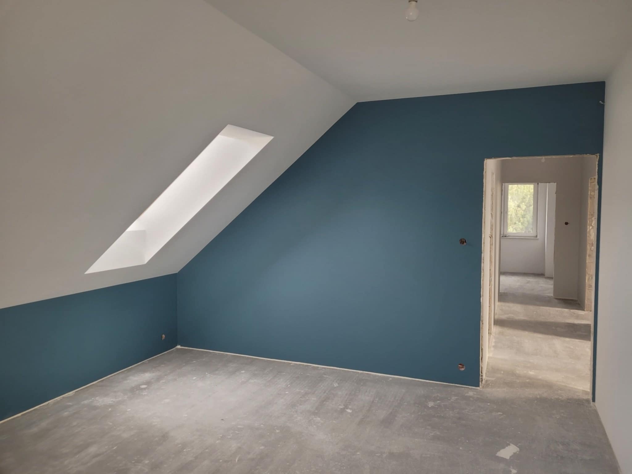 Pokój na poddaszu po szpachlowaniu, z pomalowaną na niebiesko ścianą do połowy wysokości, surową betonową podłogą i widocznym oknem dachowym oraz przejściem do kolejnego pomieszczenia.