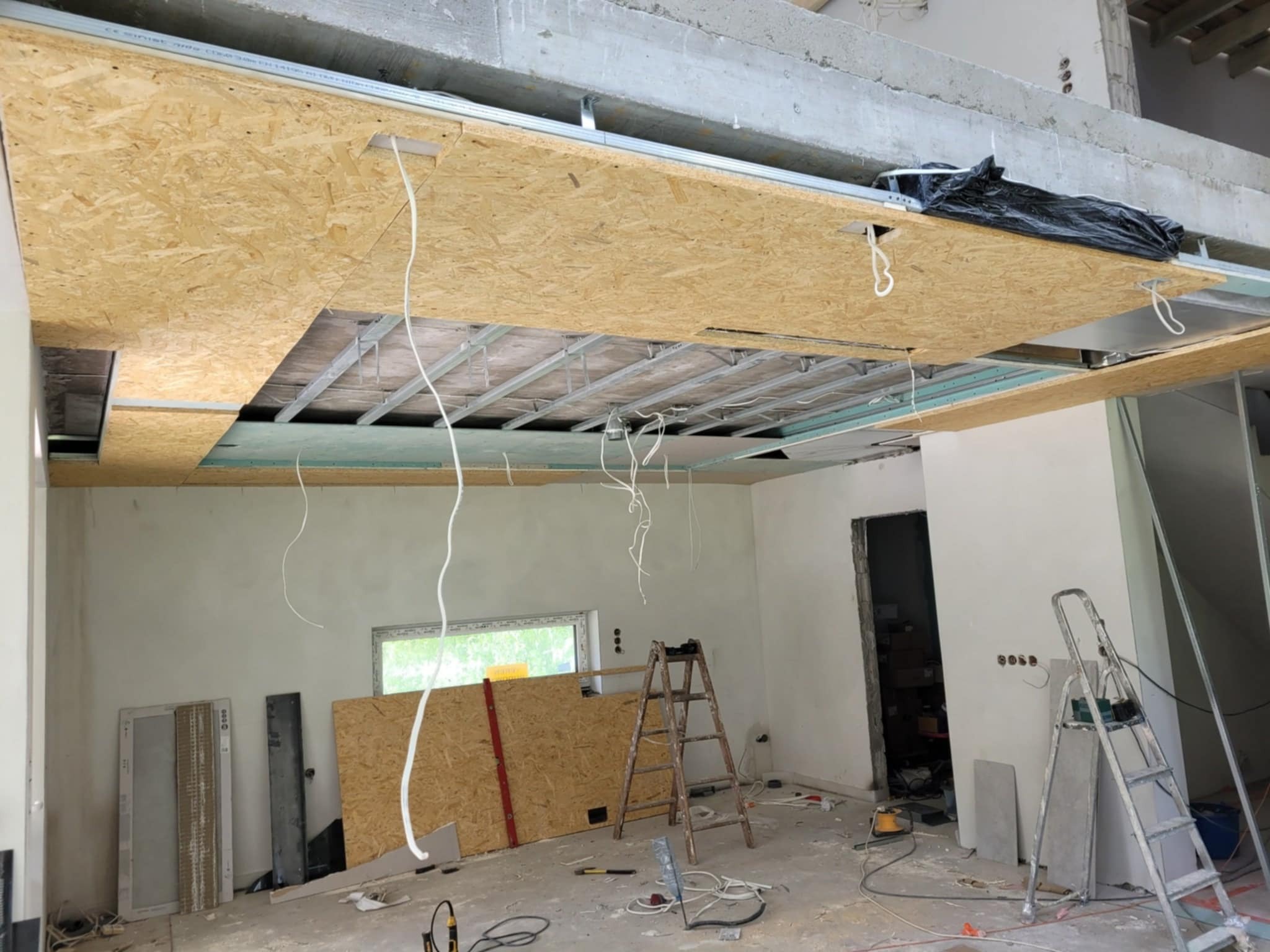 Montaż sufitu podwieszanego z płyt OSB na metalowej konstrukcji, widoczne przewody elektryczne, drabiny i materiały budowlane w surowym wnętrzu.