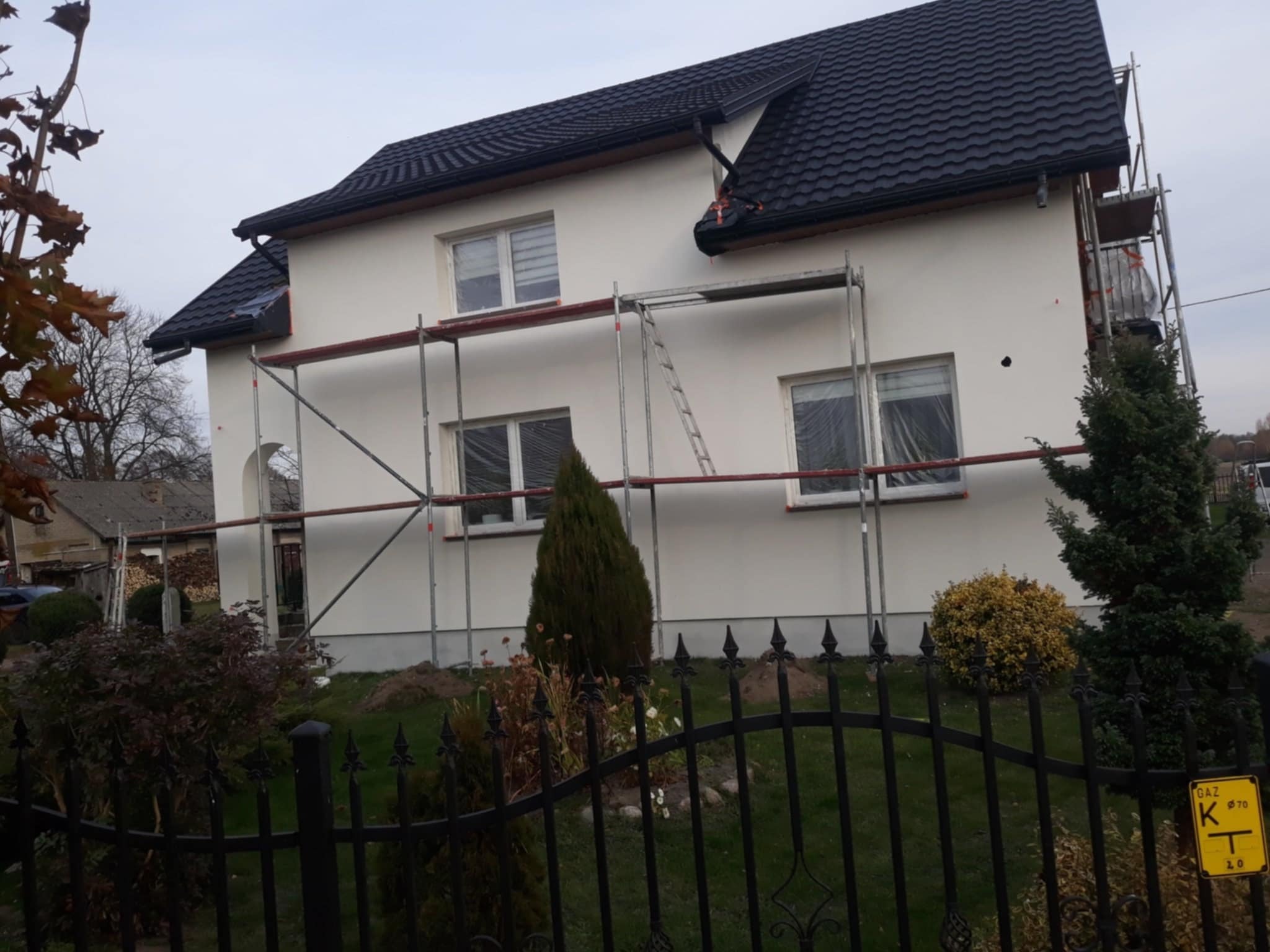 Dom jednorodzinny w trakcie prac elewacyjnych, widoczne rusztowania, okna zabezpieczone folią, świeżo nałożony tynk w kolorze złamanej bieli, czarny dach z dachówki ceramicznej, metalowe ogrodzenie...