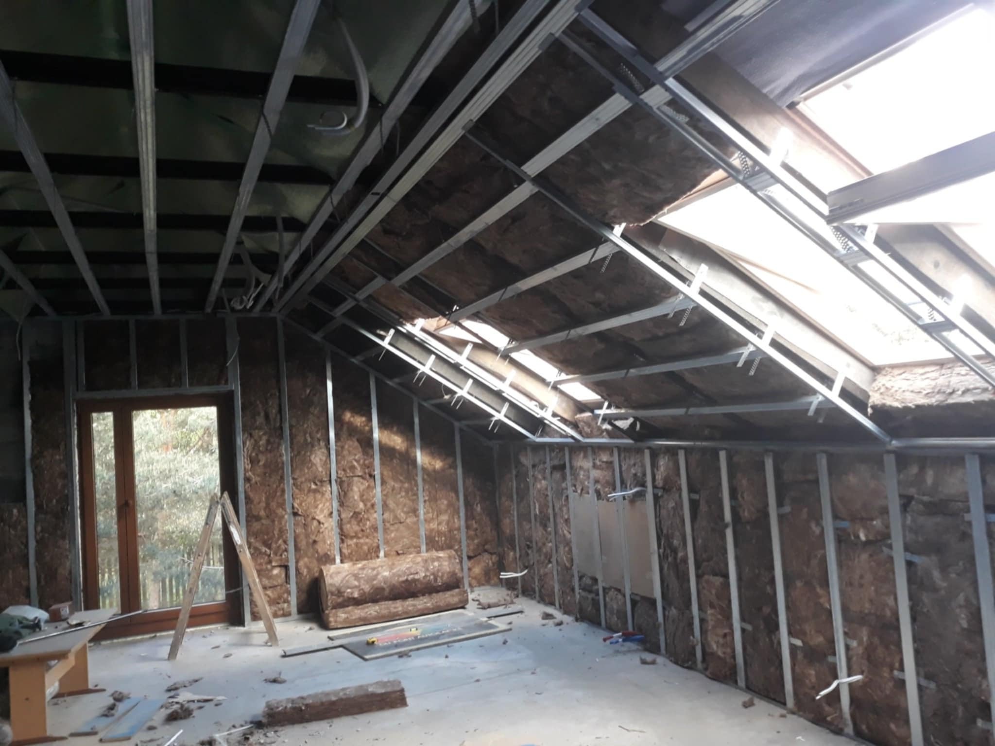 Wnętrze poddasza w trakcie prac: widoczne metalowe profile konstrukcyjne, izolacja ścian wełną mineralną, okno dachowe, częściowo zamontowane płyty na suficie, oraz rozstawiona drabina oparta o okno.
