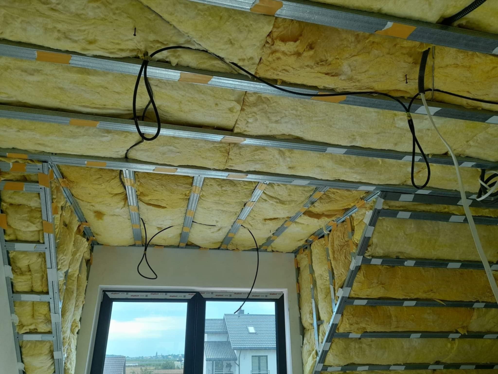 Skośny sufit poddasza w trakcie izolacji wełną mineralną, widoczne metalowe profile i przewody elektryczne, okno dachowe z widokiem na zabudowę jednorodzinną.