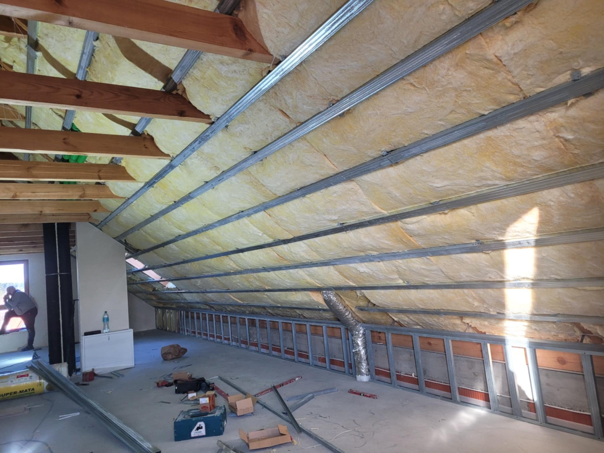 Poddasze w trakcie izolacji: widoczne metalowe profile konstrukcyjne, wełna mineralna między nimi, fragment okna z sylwetką osoby, narzędzia i materiały budowlane na podłodze.