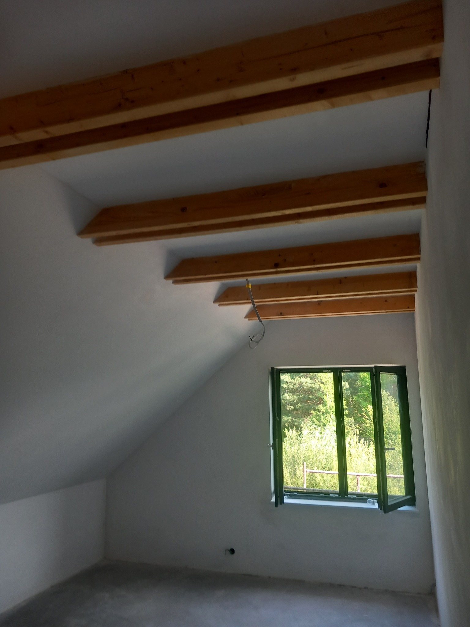 Poddasze po szpachlowaniu, z widocznymi drewnianymi belkami stropowymi i oknem z zielonym widokiem. Ściany gładkie, przygotowane do dalszych prac wykończeniowych.