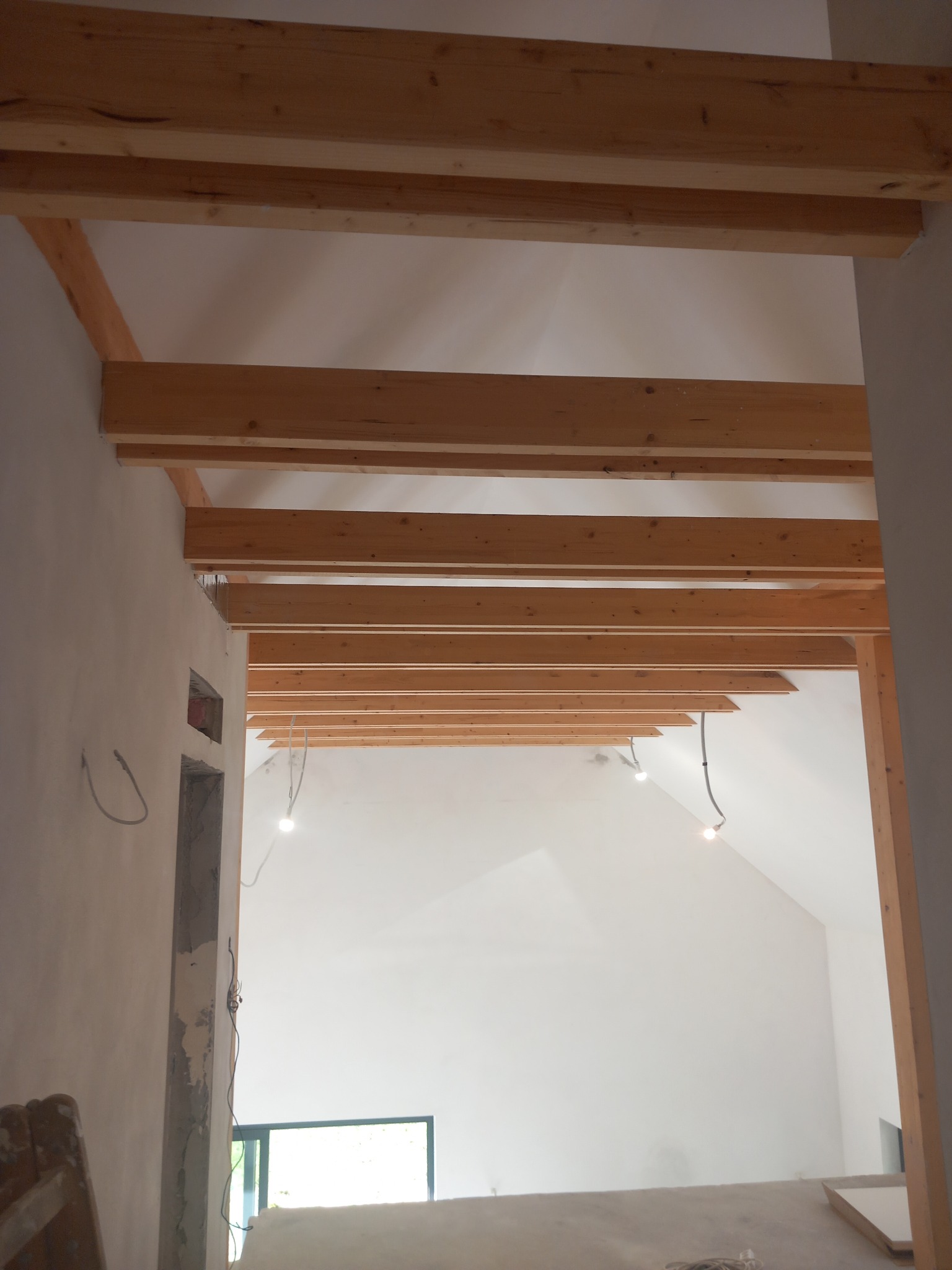 Wnętrze w trakcie prac wykończeniowych: drewniane belki stropowe, surowe ściany, widoczne przewody elektryczne. Ujęcie z dołu, perspektywa podkreśla wysokość pomieszczenia.