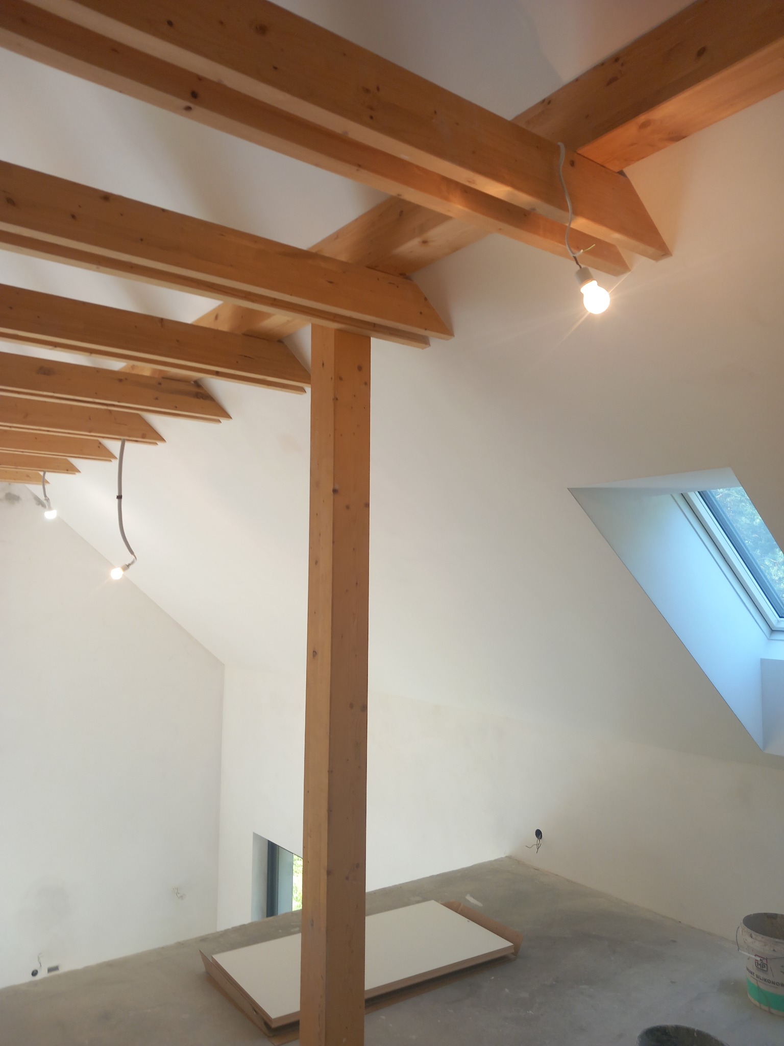 Poddasze z drewnianą konstrukcją stropu i oknem dachowym. Widoczne przygotowania do prac wykończeniowych, surowy beton na podłodze, białe ściany i elementy instalacji.