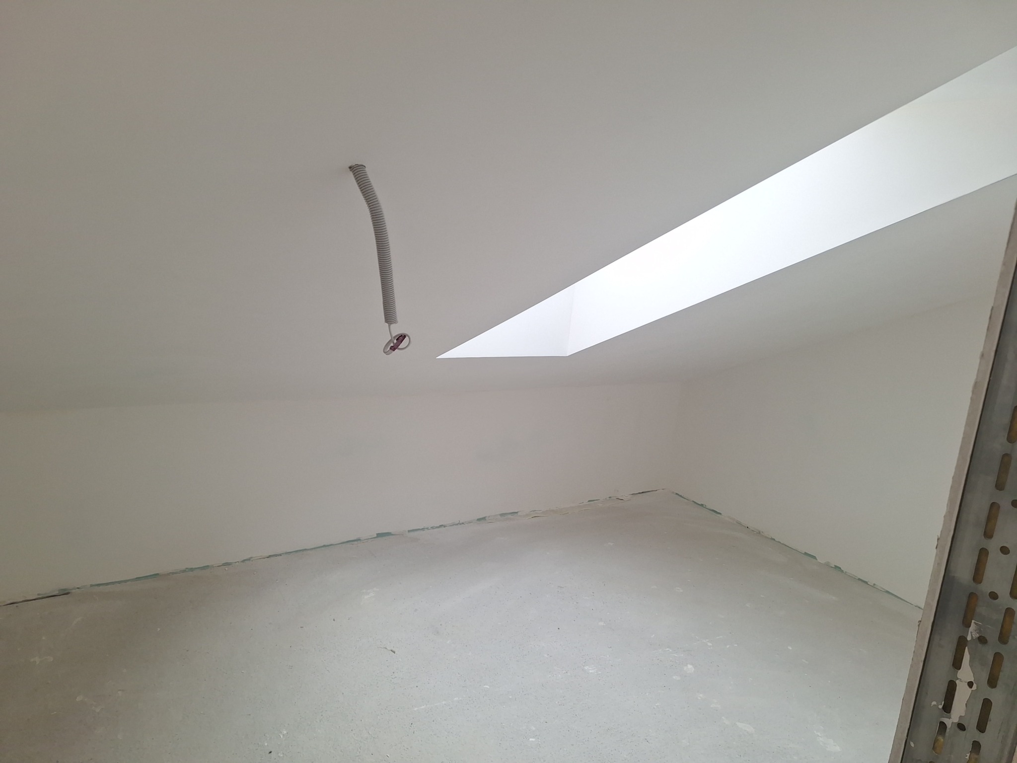 Wnętrze pokoju po szpachlowaniu, widoczne gładkie, białe ściany i sufit z oknem dachowym. Na suficie zwisa kabel w peszlu. Podłoga przygotowana do dalszych prac.