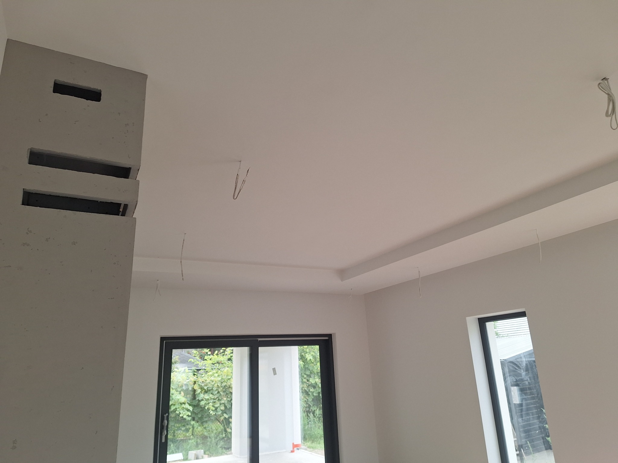 Wnętrze po gładziowaniu i szpachlowaniu, widoczne okna, sufit z dekoracyjnym elementem i fragment ściany z wnękami. Wystające kable elektryczne.