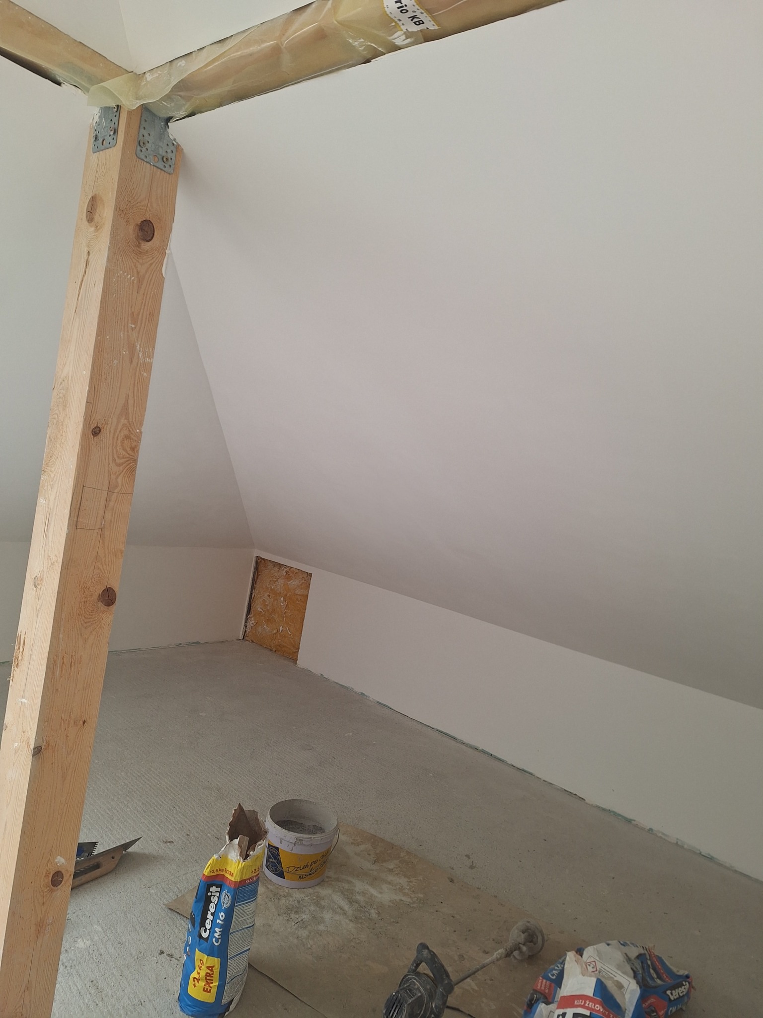Poddasze po szpachlowaniu i malowaniu, widoczna drewniana belka wzmacniająca, narzędzia i materiały budowlane na podłodze. Świeżo wykończone ściany i sufit.