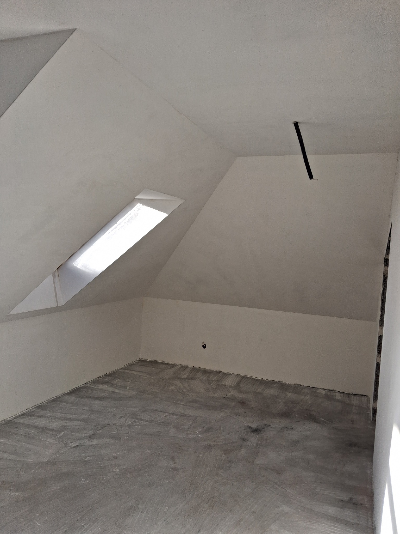 Poddasze po szpachlowaniu, przygotowane do dalszych prac wykończeniowych. Widoczne okno dachowe i wystający przewód. Szare podłoże pod panele.