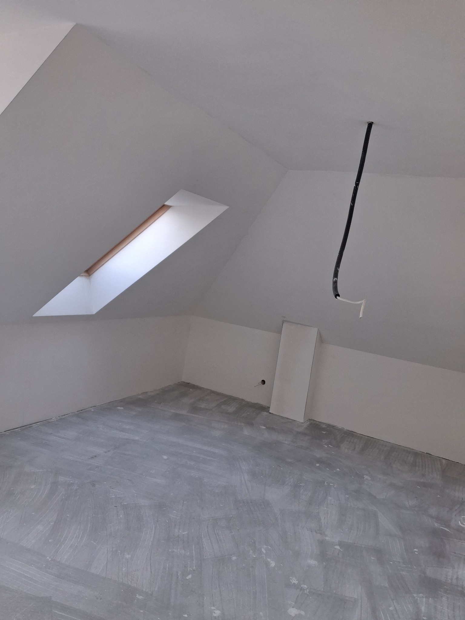 Poddasze po szpachlowaniu, przygotowane do dalszych prac wykończeniowych. Widoczne okno dachowe i wystające przewody elektryczne. Szare podłoże.